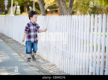 Cute Young Mixed Race Boy Walking Stick avec le long de clôture blanche. Banque D'Images