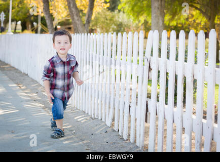 Cute Young Mixed Race Boy Walking Stick avec le long de clôture blanche. Banque D'Images