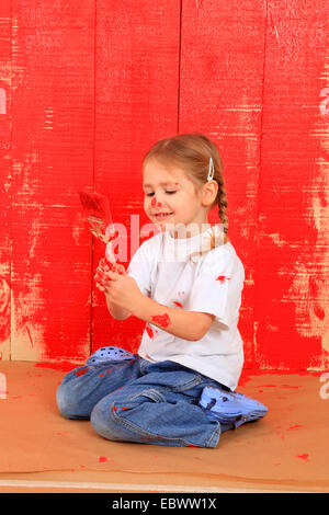 Petite fille avec un pinceau de genou en face d'un mur en bois peint rouge Banque D'Images