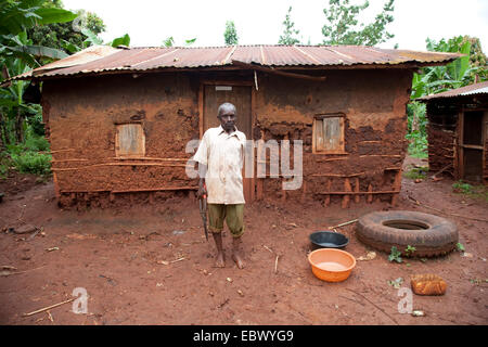 Un homme âgé est debout à l'avant de sa maison de terre simple avec un toit en tôle ondulée, l'Ouganda, Jinja Banque D'Images