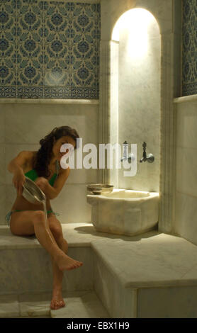 Jeune femme assise sur un banc dans un marmorn hammam verser de l'eau sur les jambes d'un metal bown Banque D'Images