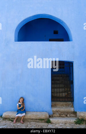 Petite fille assise sur un banc en pierre de manger des glaces devant une maison peintes en bleu, Italie, Sardaigne, Posada Banque D'Images