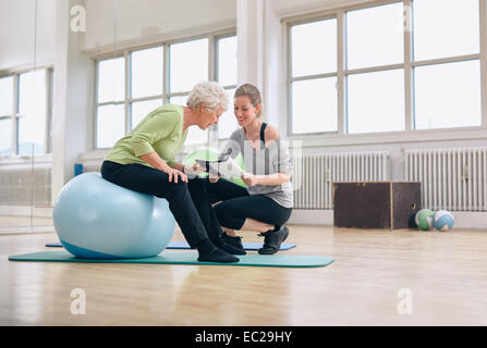 Femme âgée dans une salle de sport assise sur boule d'exercice et de parler à ses renseignements personnels formateur féminin sur un plan d'entraînement. Senior woman et Banque D'Images