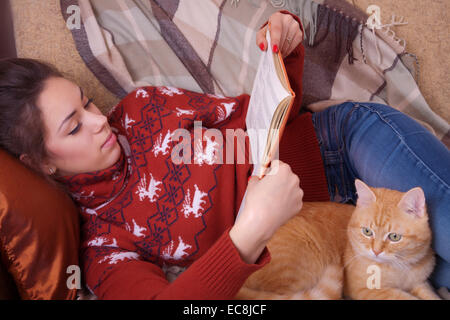 Cute girl allongé sur le canapé avec un chat rouge et la lecture d'un livre Banque D'Images