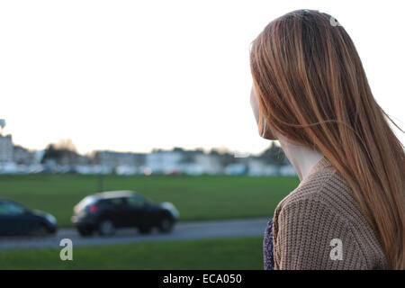 Vue arrière plan sur une jeune femme assise sur un banc, à la recherche dans la distance dans un parc avec des voitures garées. Banque D'Images