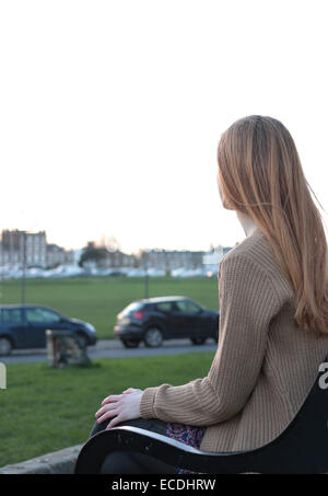 Vue arrière plan sur une jeune femme assise sur un banc, à la recherche dans la distance dans un parc avec des voitures garées. Banque D'Images