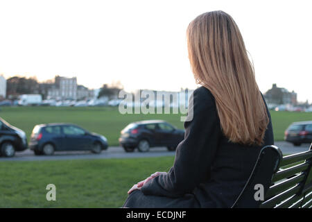 Vue arrière plan sur une jeune femme assise sur un banc, à la recherche dans la distance dans un parc avec des voitures garées. Banque D'Images