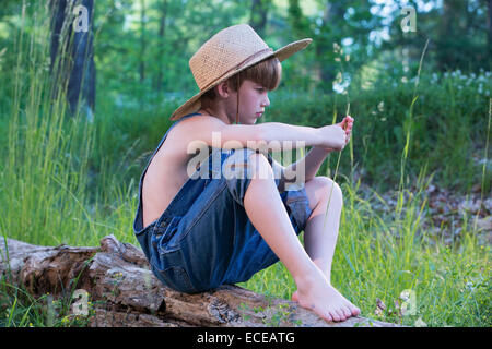 Young boy wearing straw hat sitting on log Banque D'Images