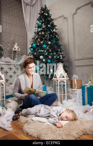 Young woman reading book pour petit garçon à côté d'arbre de Noël, boy s'endormir Banque D'Images