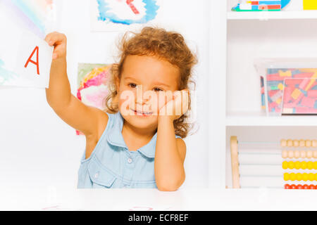 Une petite fille d'âge préscolaire avec des cheveux bouclés montrant flashcard avec une lettre dans la classe de maternelle avec calme expression positive Banque D'Images