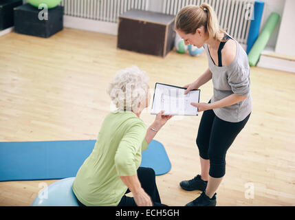 Dans une salle de gym Senior woman sitting on exercise ball et parler à son entraîneur privé sur un plan d'entraînement Banque D'Images