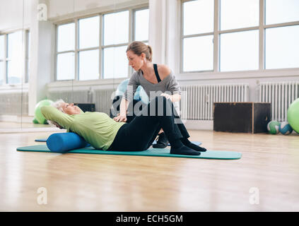 Senior woman effectuant les exercices du dos sur un rouleau en mousse étant assisté par son entraîneur personnel à la salle de sport. Thérapeute physique aidant Banque D'Images