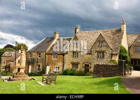 Un village vert avec un banc et une rangée de cottages en pierre de Cotswold et un ciel d'un noir d'encre. Banque D'Images
