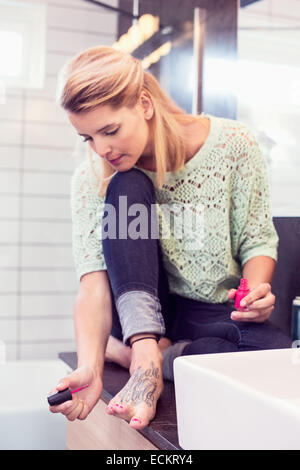 Toute la longueur d'ongles peinture jeune femme assis sur compteur de salle de bains Banque D'Images