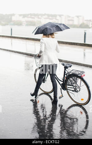 Vue arrière sur toute la longueur de businesswoman walking with bicycle on city street pendant la saison des pluies Banque D'Images