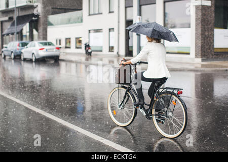 Vue arrière sur toute la longueur de businesswoman riding bicycle on city street humide pendant la saison des pluies Banque D'Images