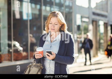 Businesswoman using mobile phone et holding Coffee cup jetable en marchant sur le trottoir Banque D'Images