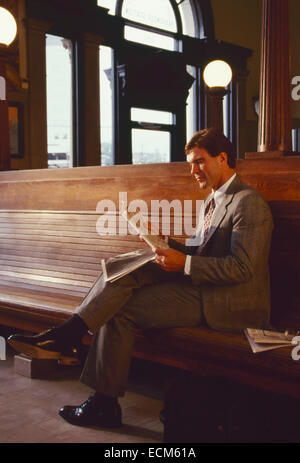 Un handsome businessman en costume lit un journal assis sur un banc en bois à une gare ferroviaire. Banque D'Images