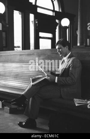 Un handsome businessman en costume lit un journal assis sur un banc en bois à une gare ferroviaire. Banque D'Images