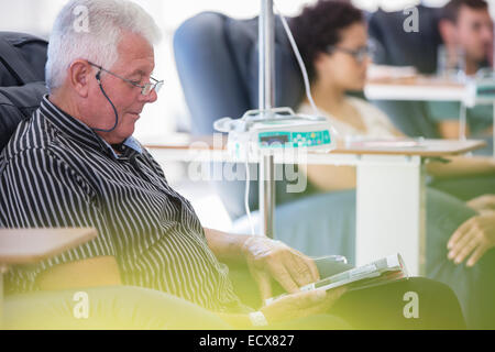 Senior patient recevant un traitement tout en restant assis et reading book in hospital ward Banque D'Images