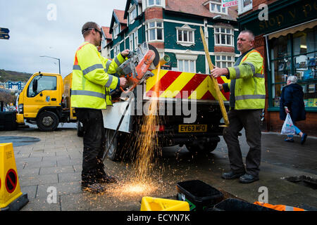 Un équipage de deux hommes du Conseil du comté de Ceredigion autorité locale du travail direct travailleurs portant hi-vis yellow jackets couper un poteau de métal avec une meuleuse d'angle de la rue, Aberystwyth Wales UK Banque D'Images