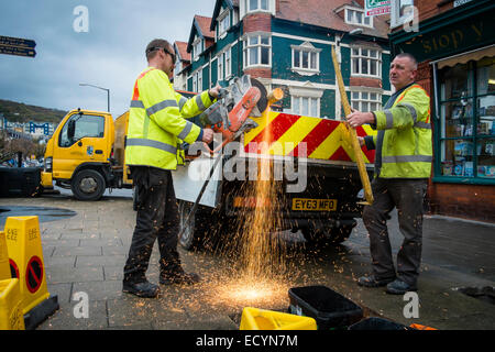 Un équipage de deux hommes du Conseil du comté de Ceredigion autorité locale du travail direct travailleurs portant hi-vis yellow jackets couper un poteau de métal à l'aide d'une meuleuse d'angle de la rue, Aberystwyth Wales UK Banque D'Images