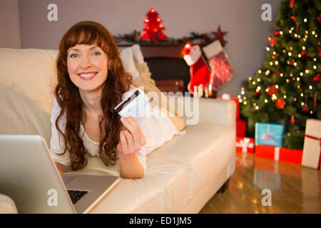 Smiling redhead lying on couch shopping online with laptop Banque D'Images