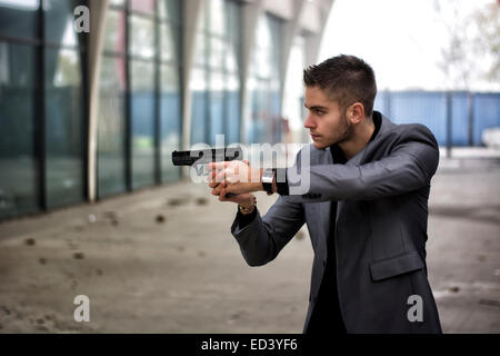 Bien habillé beau jeune détective ou policier ou gangster debout dans un environnement urbain visant une arme à feu sur la gauche Banque D'Images