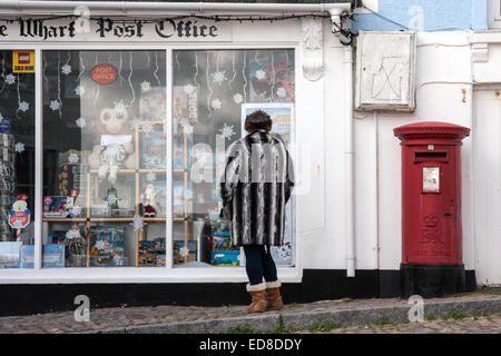 Une femme dans un manteau en fausse fourrure rayée à la recherche dans la fenêtre d'un bureau de poste auxiliaire traditionnelle à St Ives, Cornwall. Banque D'Images