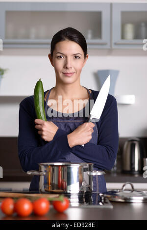 Happy young woman posing dans sa cuisine avec les bras croisés tenant un couteau et concombre comme elle prépare un diner en bonne santé Banque D'Images