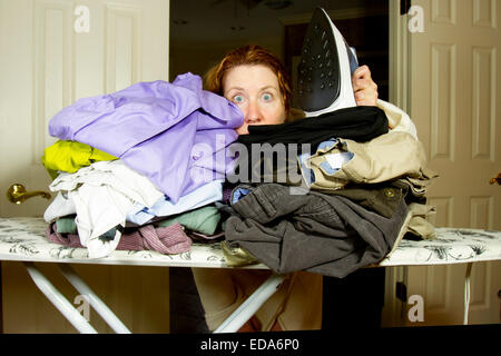 Woman peering dans un tas de vêtements unironed tenant sur une planche à repasser un fer à vapeur Banque D'Images