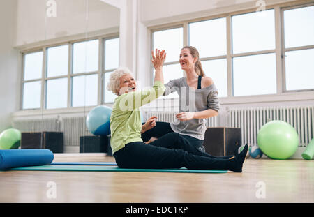 Senior woman sitting on fitness mat haute donnant cinq à son entraîneur personnel à la salle de sport. Vieille Femme excité de se réjouir de la santé la réussite Banque D'Images