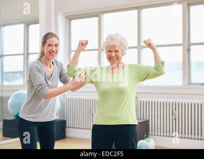 Portrait d'un bon vieux woman flexing ses bras et montrant ses muscles avec un entraîneur personnel at gym Banque D'Images