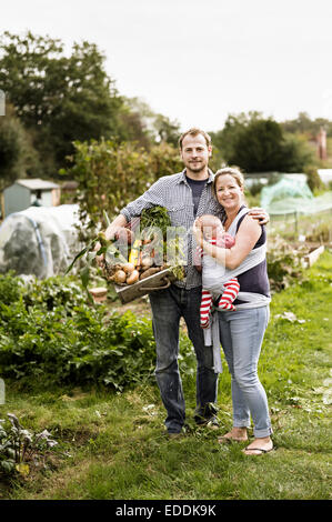 Jeune famille debout dans leur attribution, souriant. Homme tenant une boîte pleine de légumes fraîchement cueillis. Banque D'Images