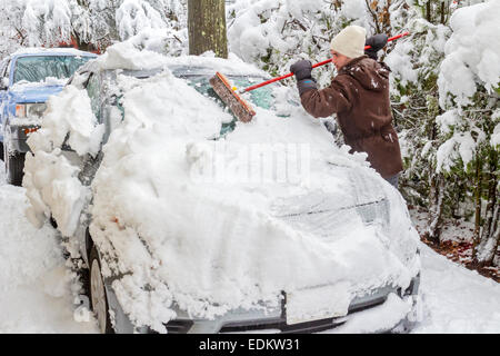 Jeune homme vêtu d'un manteau d'hiver, chapeau et gants nettoie la neige de voiture après tempête à l'aide d'un pinceau. Banque D'Images