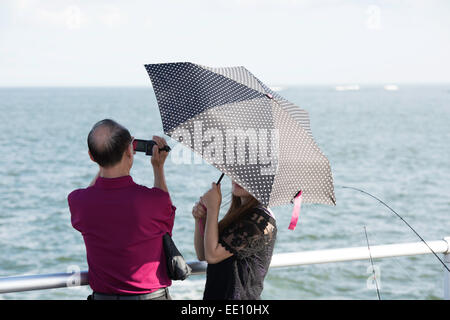 Vue arrière de l'older Asian man avec caméscope et la femme cachée sous un polk-a-dot umbrella debout à une rambarde regardant l'océan Atlantique. Banque D'Images