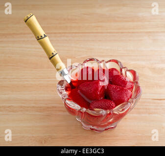 Un petit bol de verre rempli de fraises en conserve dans un liquide avec une cuillère insérée dans le fruit au-dessus d'une table en bois. Banque D'Images