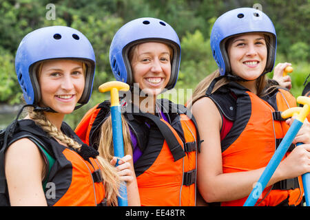 Trois jeunes femmes se tiennent côte à côte dehors, souriant à la caméra. Ils portent des casques bleus et des gilets de sauvetage orange vif avec des bretelles noires. Chaque femme tient une pagaie bleue avec un Banque D'Images