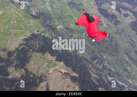 Un pilote wingsuit dans son costume est birdman rouge volant au-dessus de champs verts. Il double ainsi son temps de vol et il sourit. Banque D'Images