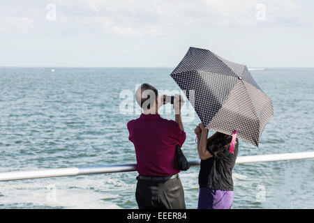 Vue arrière de l'older Asian man avec caméscope et la femme cachée sous un polk-a-dot umbrella debout à une rambarde regardant l'océan Atlantique. Banque D'Images