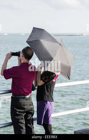 Vue arrière de l'older Asian man avec caméscope et la femme cachée sous un polk-a-dot umbrella debout à une rambarde regardant l'océan Atlantique. Banque D'Images