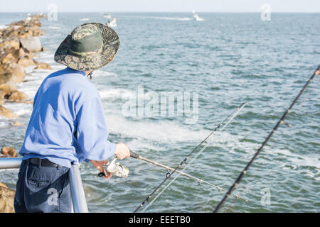 Homme avec chapeau de soleil vu de dos à la balustrade avec canne à pêche dans la main en voyant l'océan Atlantique, la pierre jetée et bateaux, côte est FL Banque D'Images