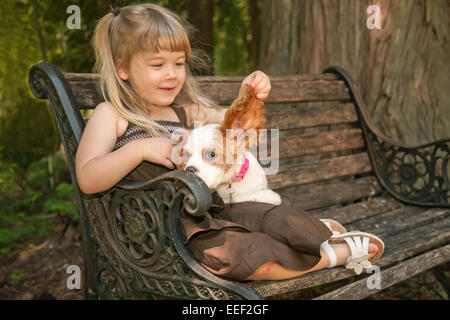 3 ans, assis sur un banc de bois, tenant l'oreille d'une 15 semaine vieux Cavalier King Charles Spaniel, Mandy Banque D'Images