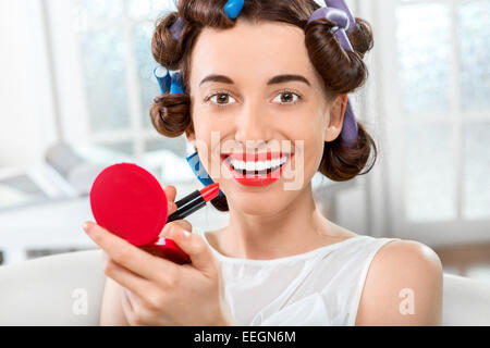 Femme souriante avec les bigoudis en utilisant du rouge à lèvres fond intérieur blanc Banque D'Images