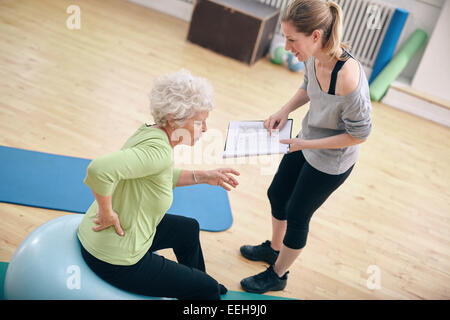 Senior woman sitting on a fitness ball avec son instructeur féminin expliquant plan d'exercice à la salle de sport. Thérapeute physique avec de vieux w Banque D'Images