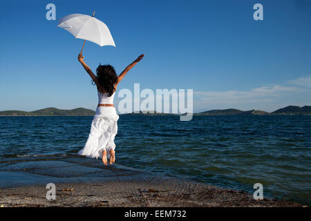 Jeune femme en blanc holding umbrella jumping on beach Banque D'Images