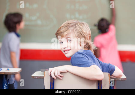 Classe élémentaire ethniques multiples. Enfant regardant à l'appareil alors que des camarades de classe de tableau. Banque D'Images