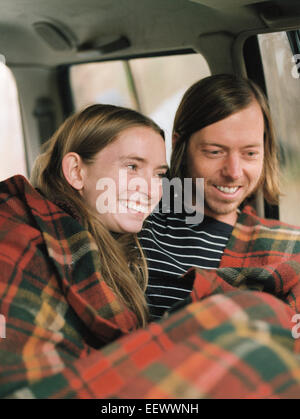 A smiling couple assis dans leur voiture, recouvert d'une couverture. Banque D'Images