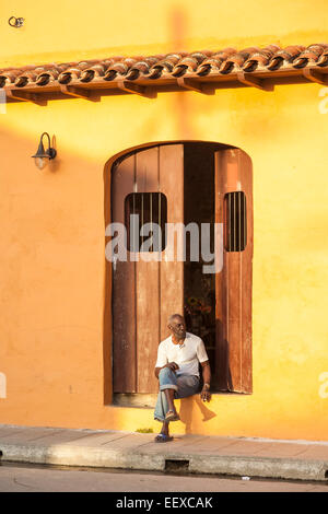 Scène de rue typique locale : un homme âgé assis dans l'embrasure d'une maison d'orange, de détente, de fumer à Camaguey, Cuba, troisième ville Banque D'Images