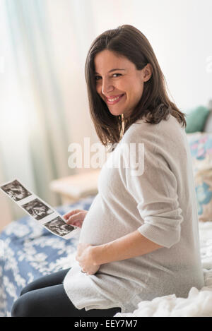Pregnant woman holding ultrasound, sitting on bed smiling Banque D'Images